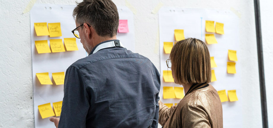 Eine Frau und ein Mann stehen vor einer Wand und kleben Post-its auf Flipchart-Papier.